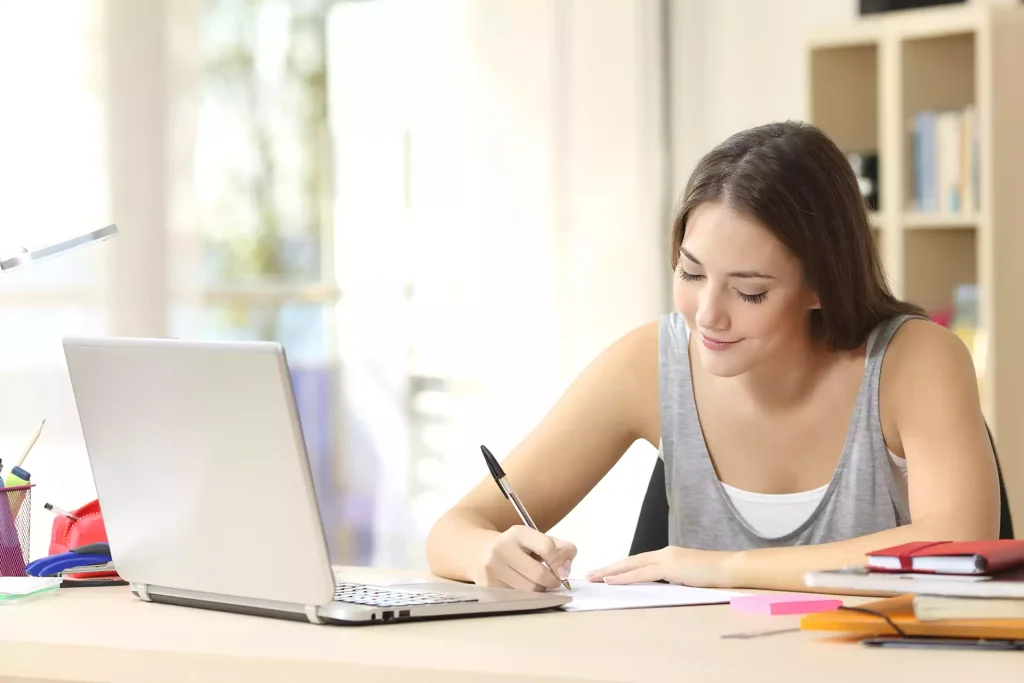 Young woman writing notes at desk with laptop in home office